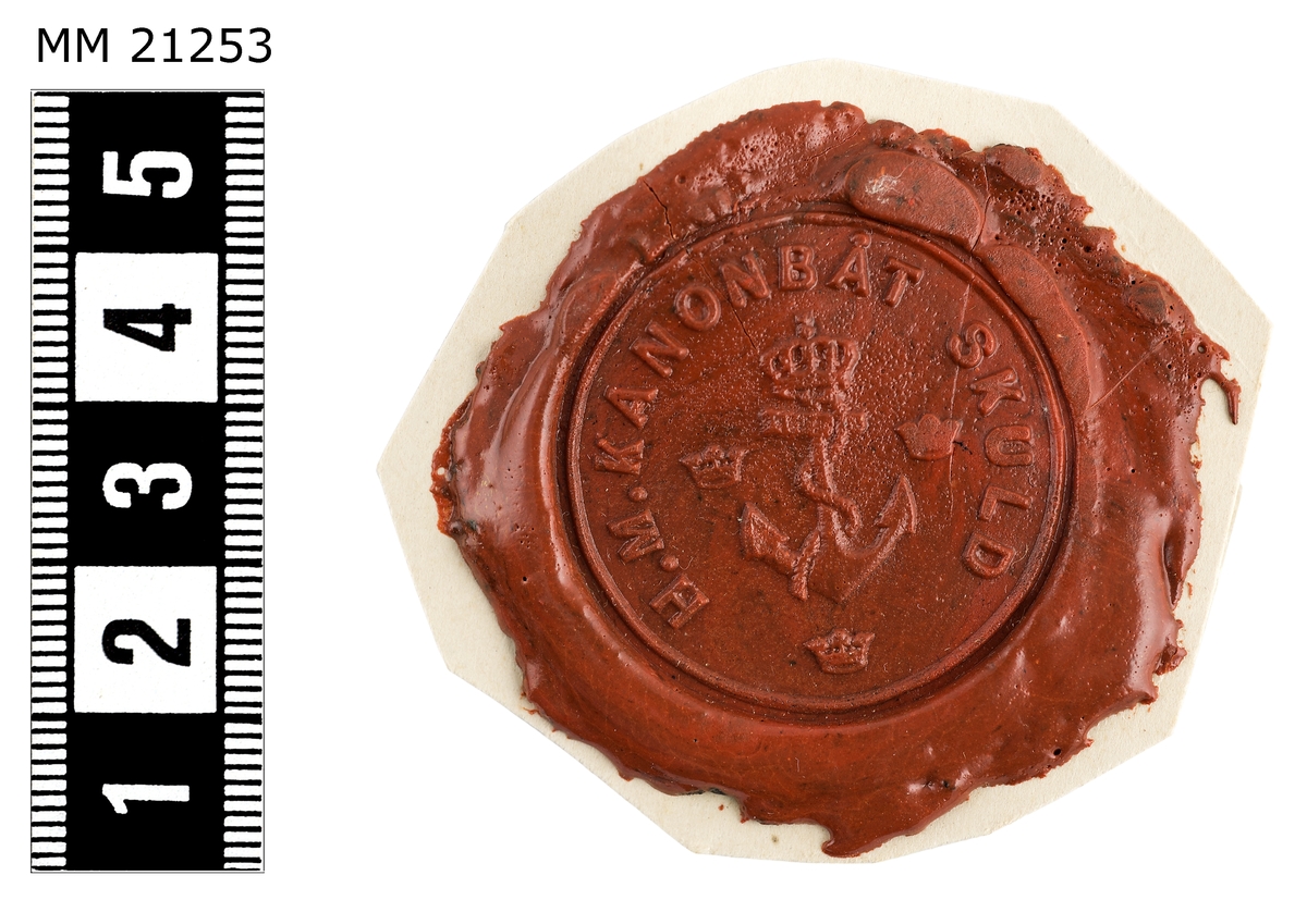Sigillavtryck av rött lack på papper. I mitten krönt stockankare omgivet av tre kronor. Längs kanten text: "H.M. kanonbåt Skuld".