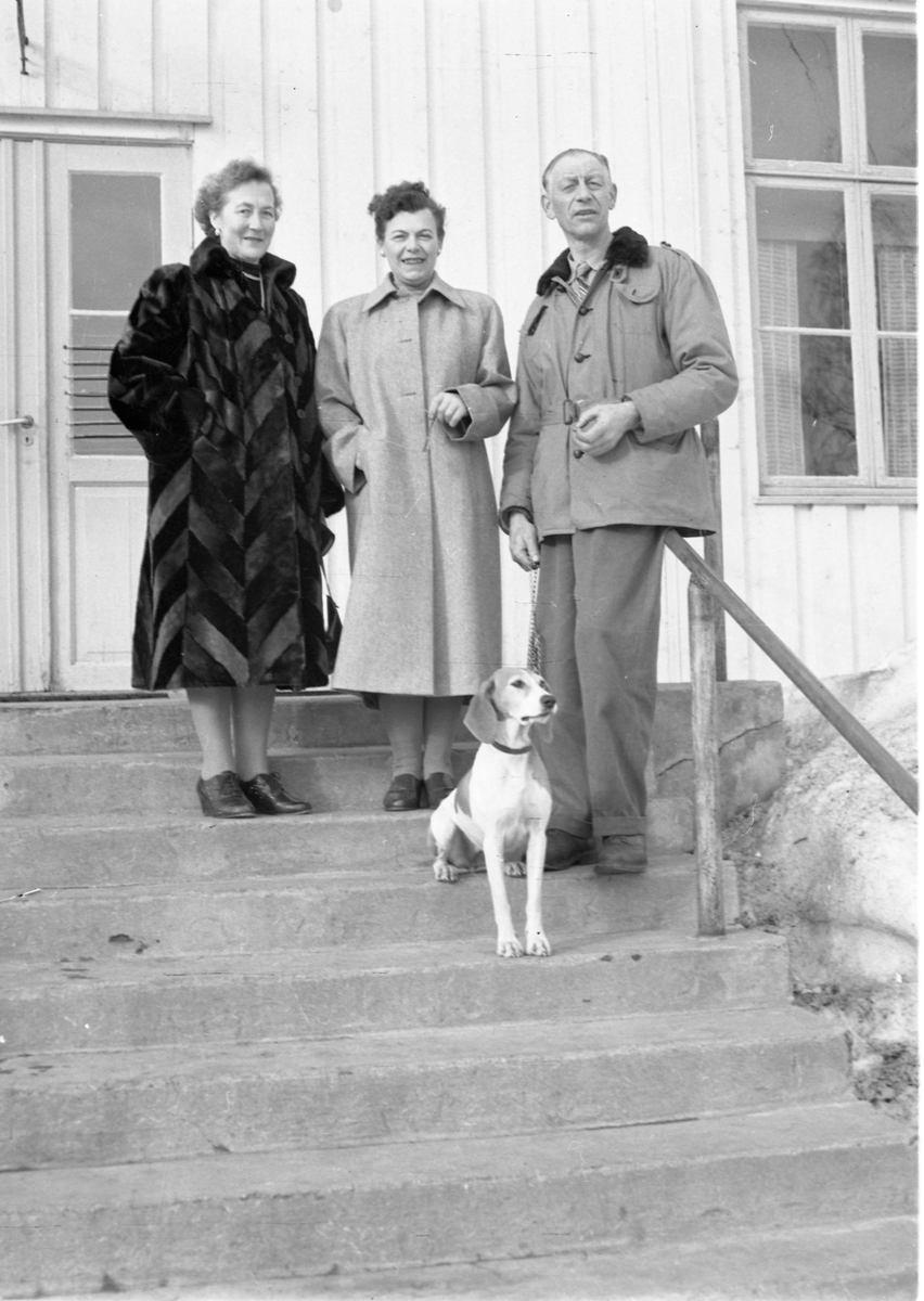 To kvinner, en mann og hund på trappen foran forsamlingshus i helfigur.