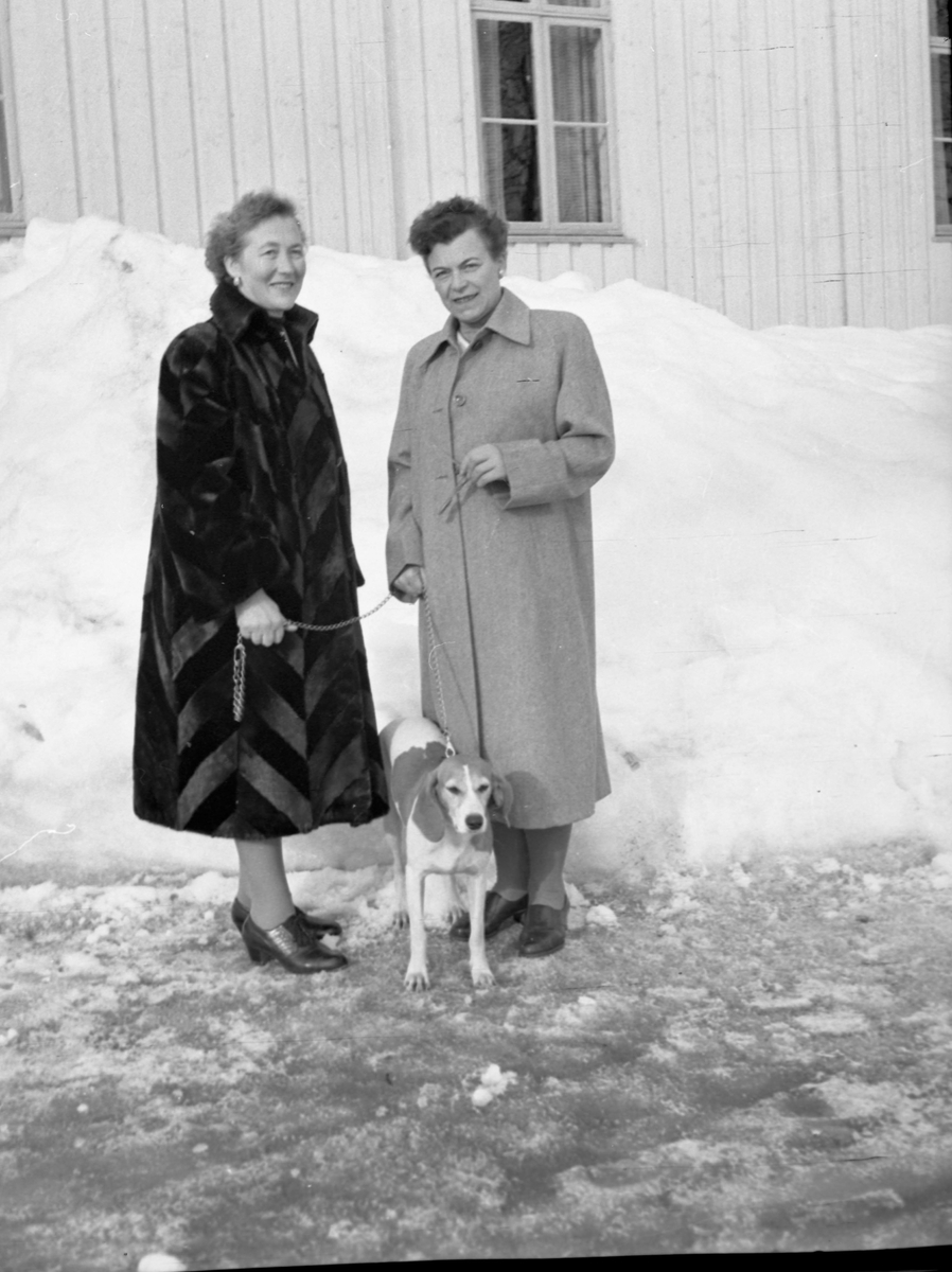 To kvinner og en hund i helfigur utendørs foran et hus