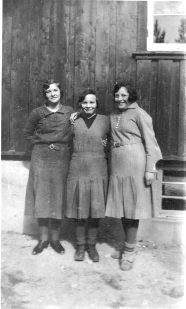 Amatørportrett av tre kvinner stående ved husvegg.