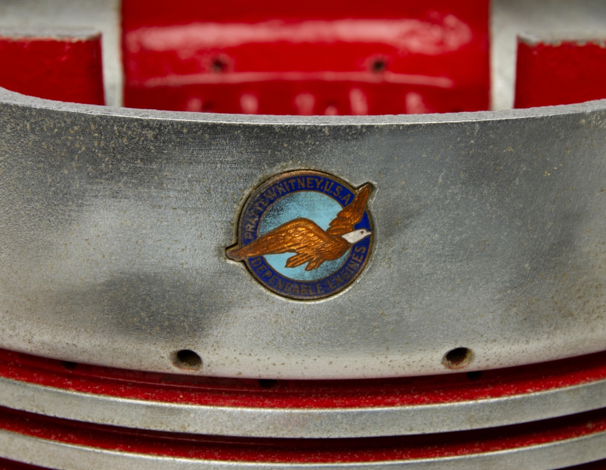 Kolvlock för flygplansmotor. På kolvlockets ena sida står det graverat "ERIK NELSON JUNE 21, 1940". På andra sidan en rund logotyp med en flygande örn i mitten och texten "Pratt & Whitney Dependable Engines". På insidan är kolvlocket målat med röd färg.