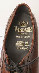 Nærbilde av brun sko med Windsorlogo med gullskrift nedi skoen.