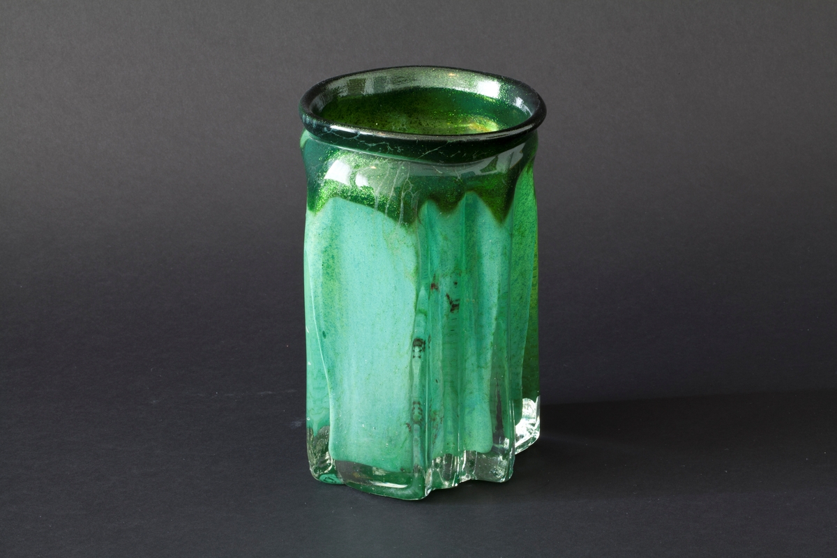 Tilnærmet sylindrisk vase i klart glass med innlagt dekor. Vasens tykke vegger har bølgende former og avsluttes i en sirkulær irregulær munningsrand. Innslag av fargede partier i grønt samt luftbobler og metallspon.