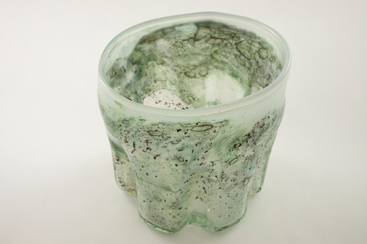 Irregulær sylinderformet vase i halvgjennomskinnelig melkehvitt glass. Ujevn munningsrand. Korpus er derkorert med metallspon, luftblærer og blå-, sort- og grønnfargede partier.