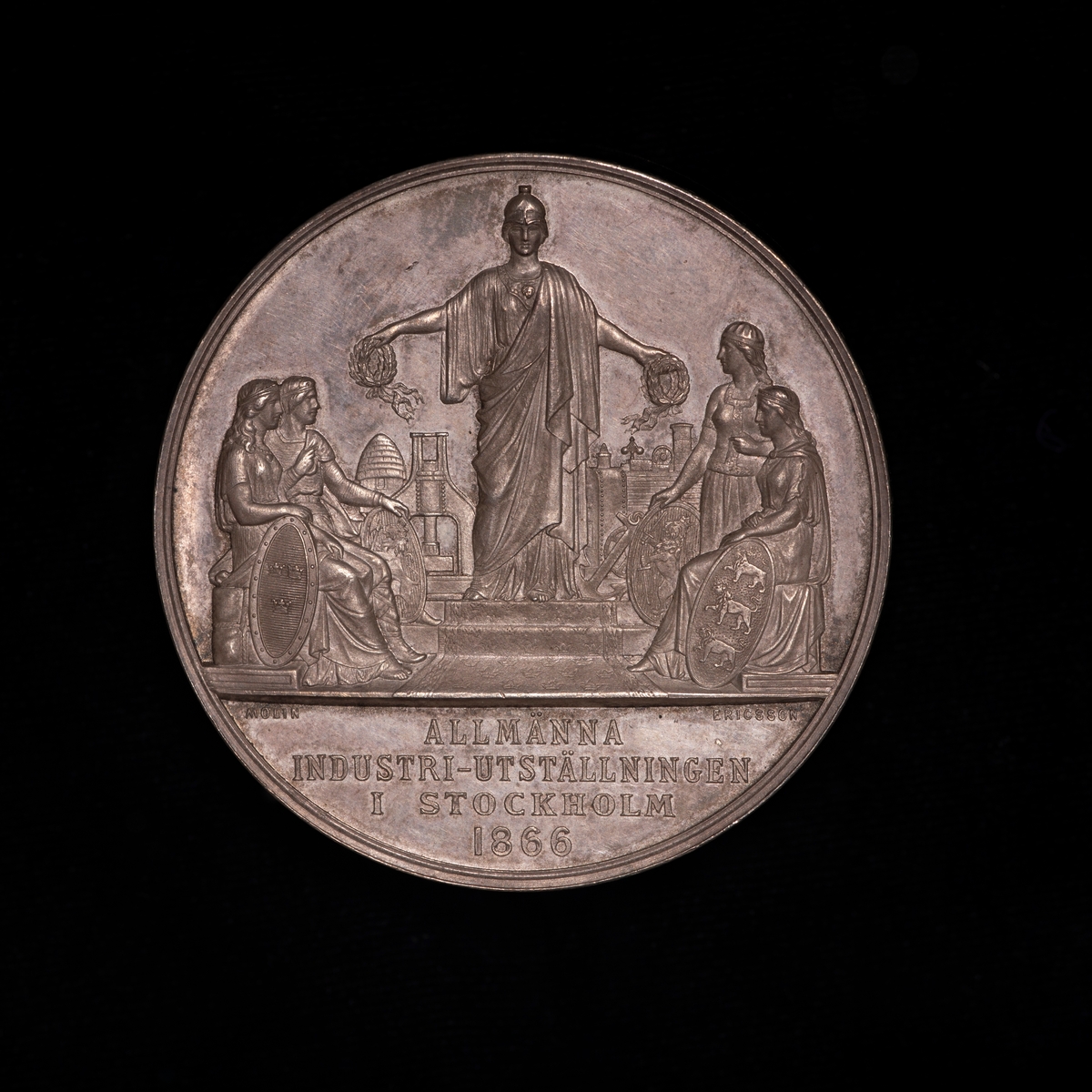 Sølvmedalje fra utstilling i Stockholm 1866. På adversen portrett mot høyre av kong Carl 15. På reversen kvinnelig guddom deler ut laurbærkranser til fire sittende kvinner.