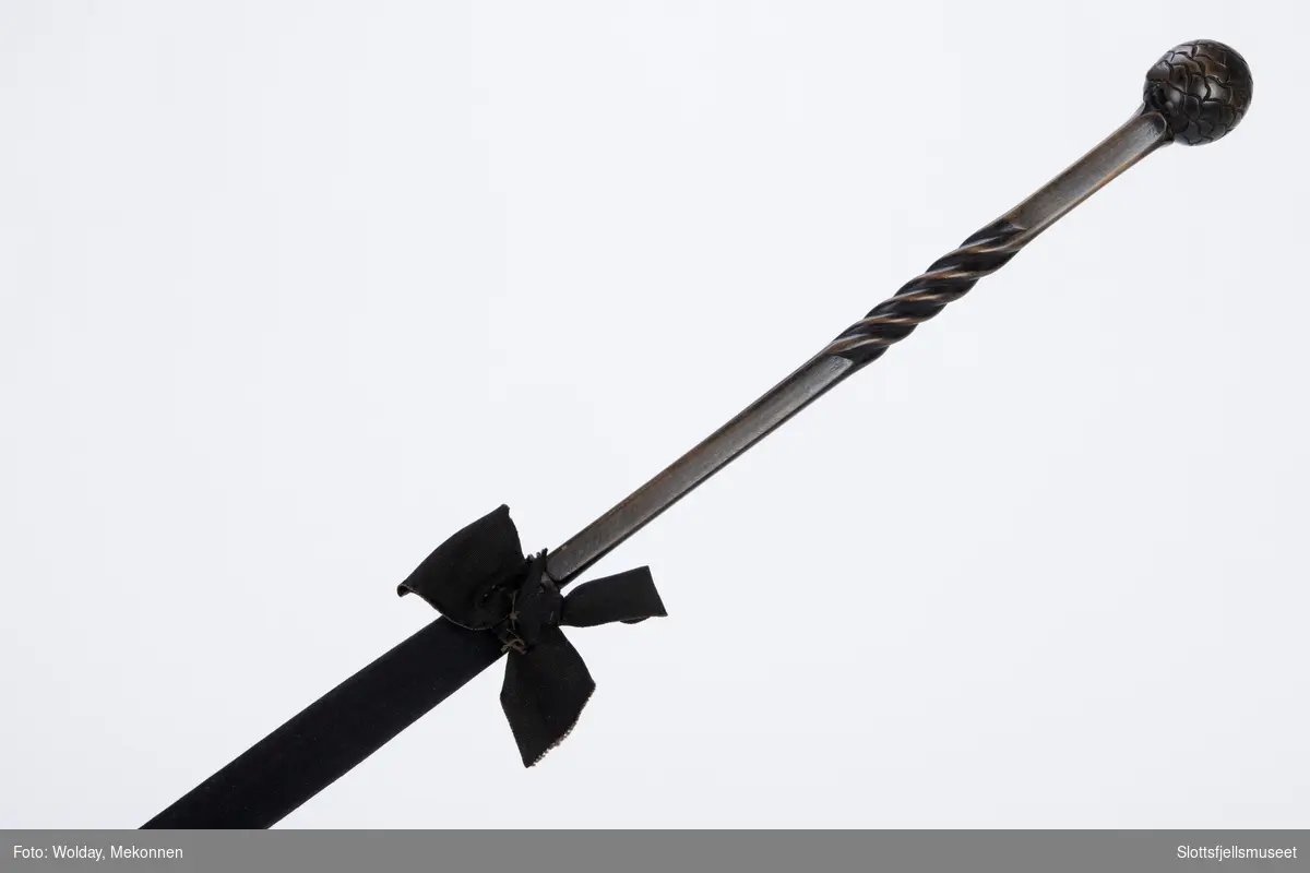 Spaserstokk - ser ut som en sammenslått paraply med svart etui av stoff. Nær håndtaket er festet en svart stoff sløyfe. Skaftet av tre som ender i en kuleformet knott.