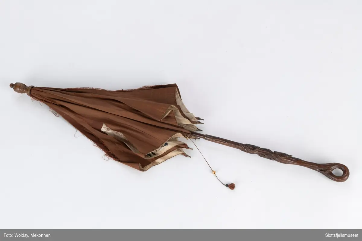 Parasoll med  8 spiler av metall. Parasoll har en utskåret treskaft og er trukket utvendig med brunt silke, innvendig med lys silke. Ved en av spilene er en snor med dusk montert for å holde spilene sammen når parasoll er lukket. 