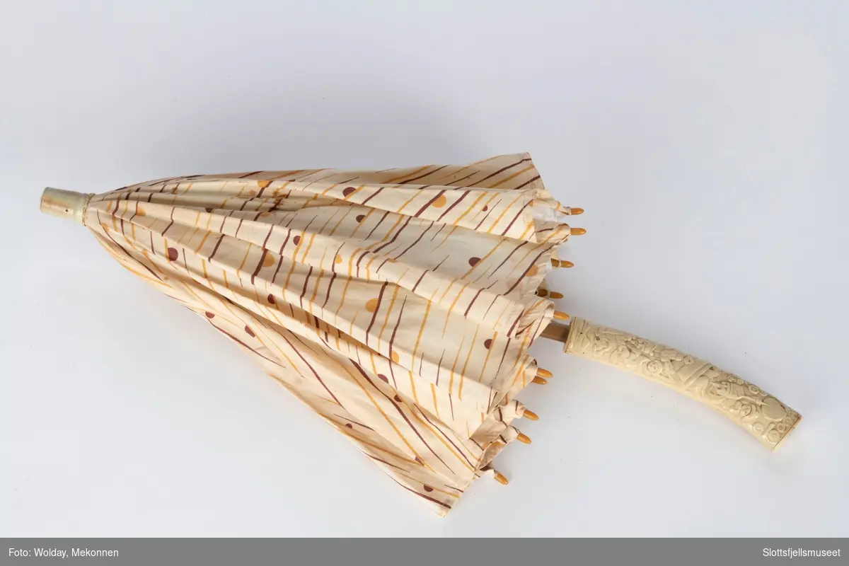 Parasoll med 12 spiler, trukket med lyst bomullsstoff. Treskaft som ender i en håndtak av bein. Svakt buet håndtak med utskjæringer i asiatisk stil. 