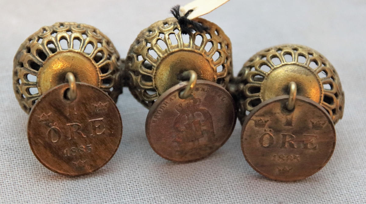 Brosch. Bestående av tre stycken i rad sittande kupolformade stycken i mässing, perforerade. Tre ettöringar är fästa på smycket med ringar, 2 st med årtalet 1885 och ett med årtal 1875.