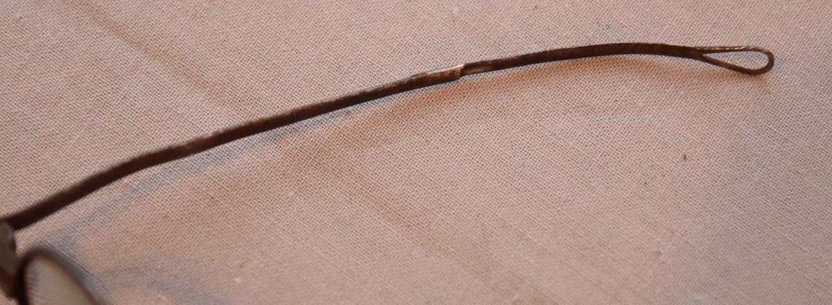 Glasögon med runda glas. Skalmarna är tillplattade och avslutas med en ögla som är avsedd för sammanbindande av ett snöre bakom huvudet. Näsbågen är skadad och lagad med en bit fastlödad ståltråd.