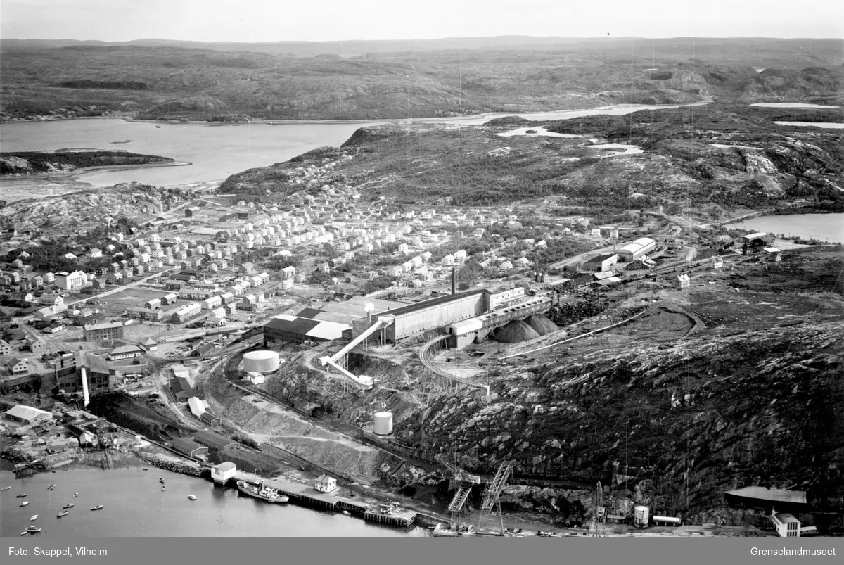 Flyfoto av Kirkenes med AS Sydvaranger med havn og andre anlegg,  ut mot Prestøya, Svartaksla og Elvenes. Til venstre i bildet ses deler av Førstevann.