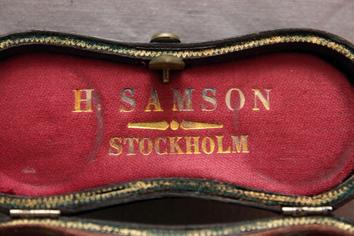 Kikare i fodral. Kikaren är utfällbar och är  gjord i mässing, ben och glas. Fodralet har en stomme av trä, är täckt på utsidan av skinn klädd i rött tyg innuti. På insidan av locket är det med guldig text tryckt "H. Samson Stockholm" (optiker från Stockholm under det sena 1800-talet). Fodralet har en låsmekanism med en liten knapp på utsidan med en inristad blomma och borrade dekorativa hål på.