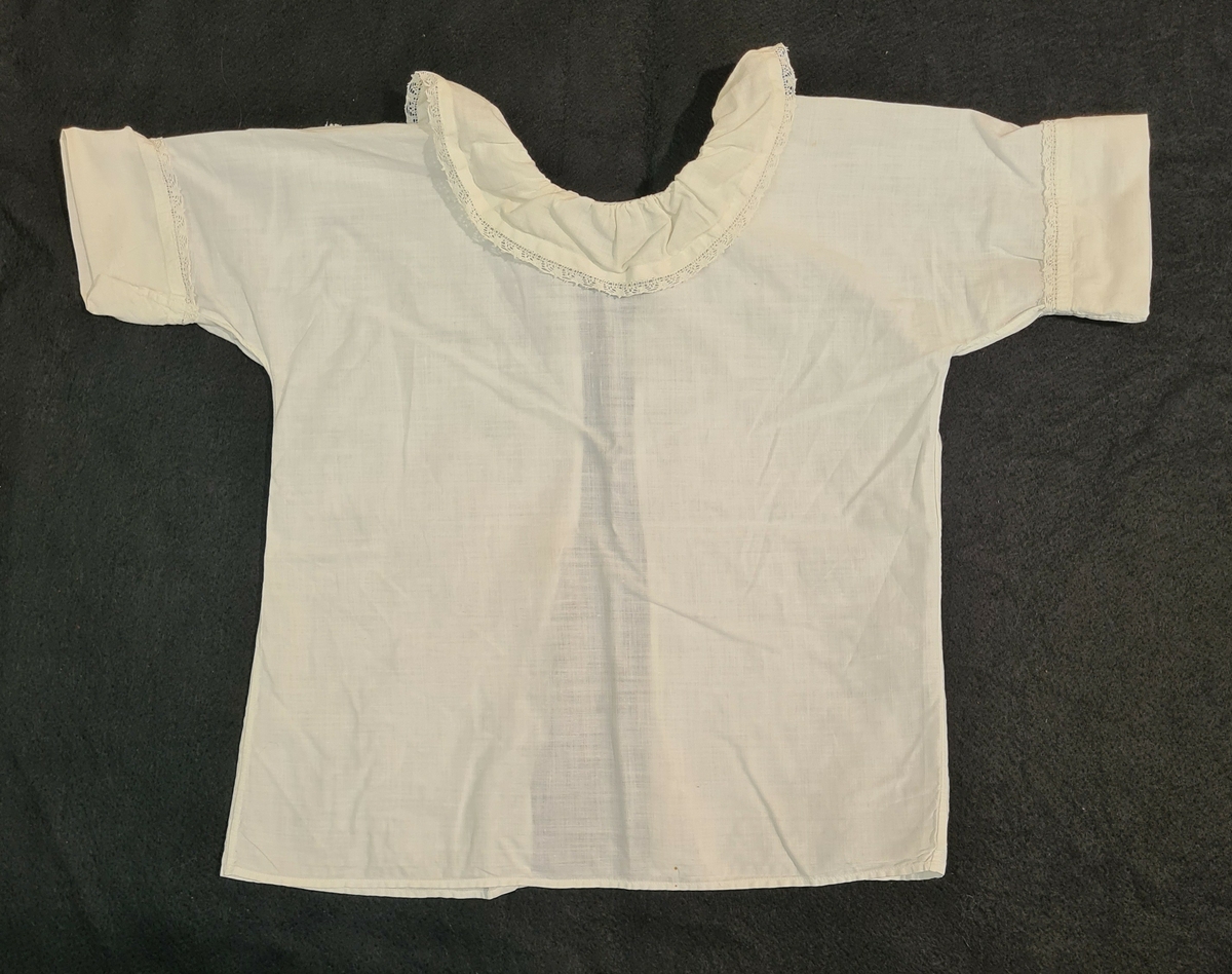 Skjorta, bomull, knypplad spets runt hals och ärmar. L. 34cm.

Ingår i en samling babykläder = mantel, blöja, stjärtlapp, gördlar och skjorta.