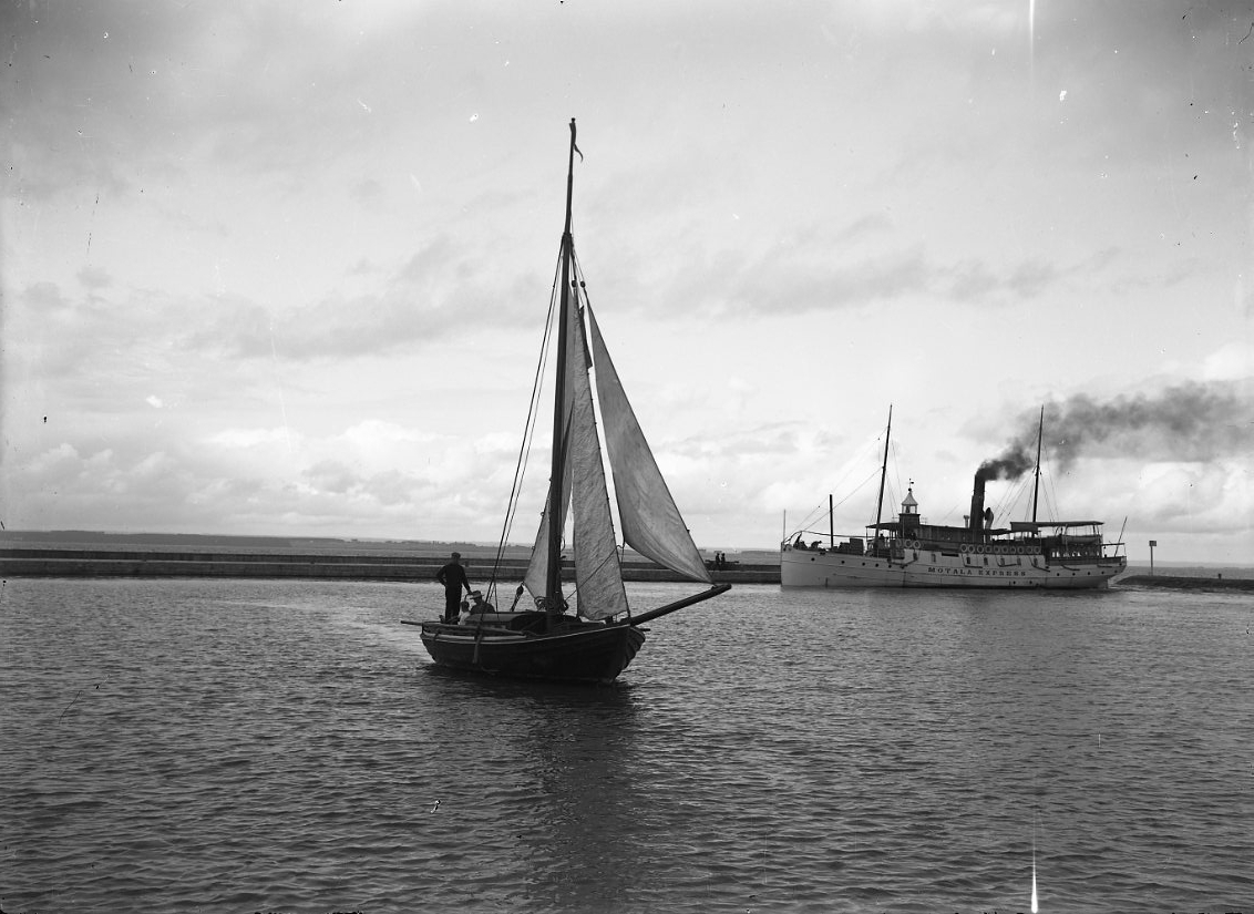 Ångbåten Motala Express ligger vid kaj i Gränna hamn, det ryker ur skorstenen. En segelbåt i förgrunden med satta segel.
