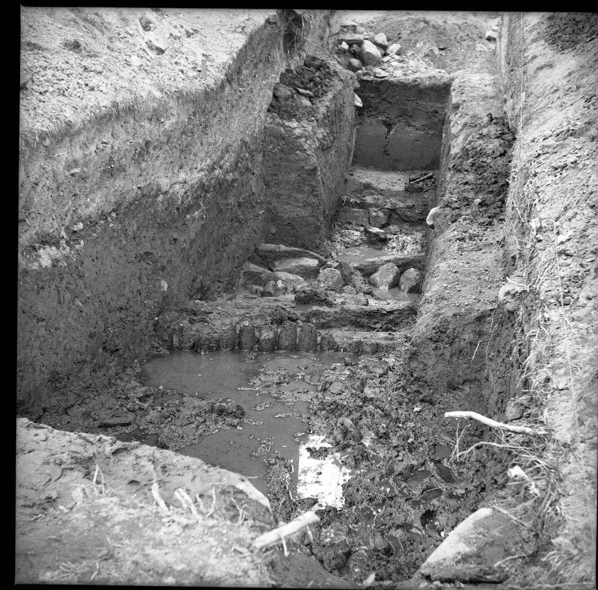 Schakt D genom södra delen av borgkullen vid Rumlaborg i Huskvarna. Fotot är taget i samband med en arkeologisk undersökning sommaren 1938. På bilden syns rester efter en palissad och bakom den stenar och andra stockrester.