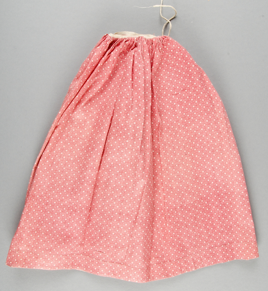 Dockklänning, blus och kjol av bomullslärft med tryckt mönster, rött med vita prickar. Livet knäpps bak, draperingar på framstycket. Spetskant.
