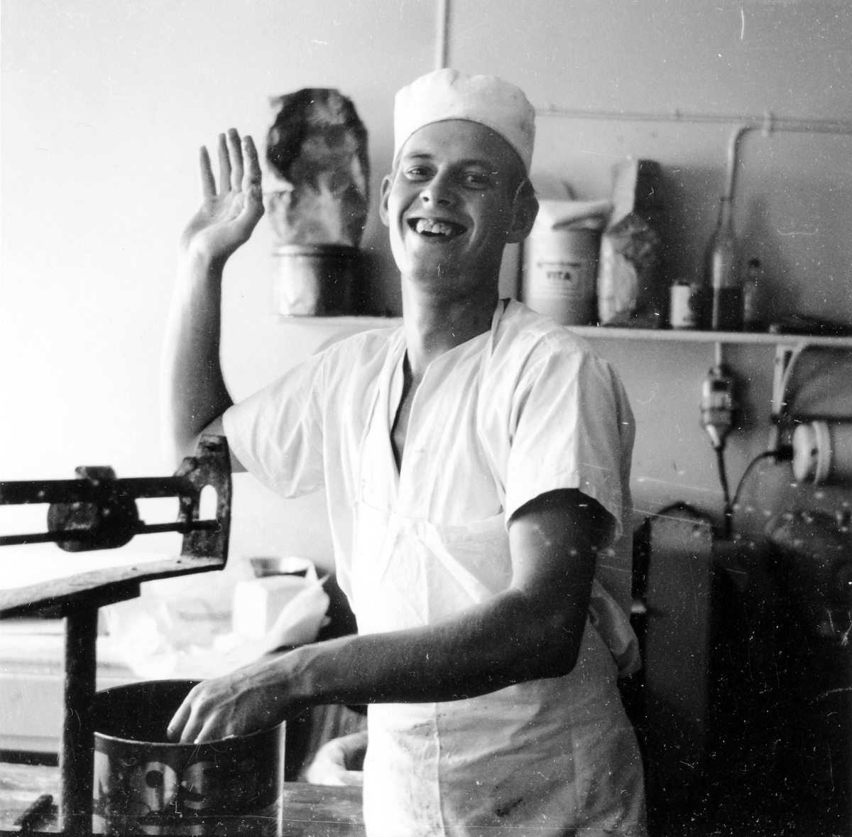 En man i vit rock och mössa vinkar leende mot kameran. Köket på Gustavs konditori, kv Gustaf vid Stora torget. 
Innehavaren av konditoriet hette Gustav Johansson enl adresskalendern 1950.
