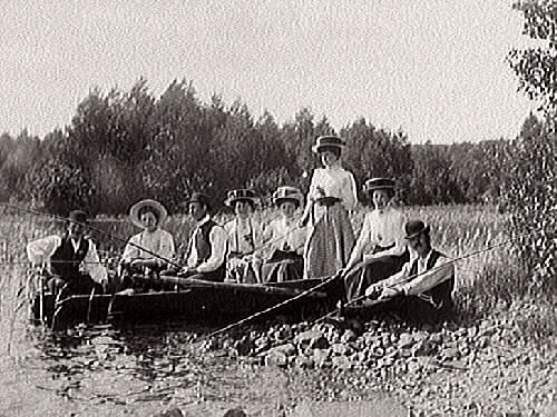 Utflykt. Tre män och fem kvinnor sitter och metar, några på stranden och några i en båt intill. Kvinnorna ser ut att bära likadana hattar.