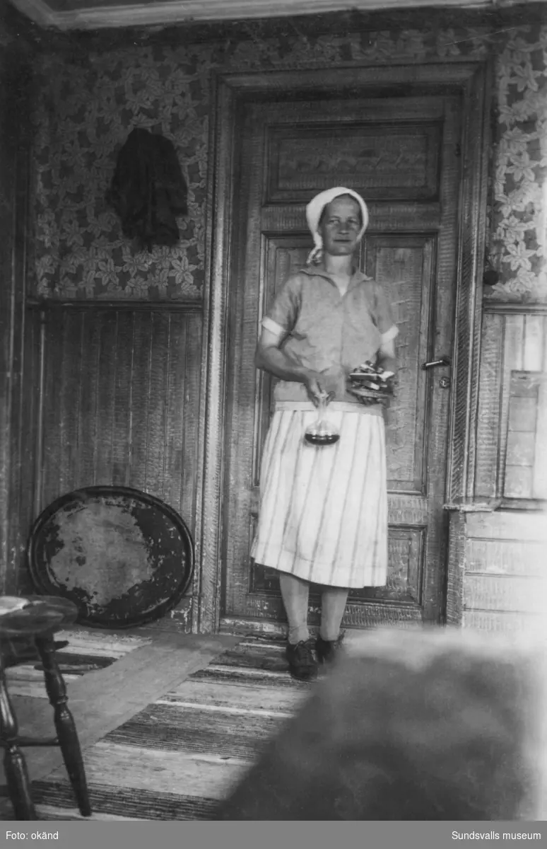En kvinna i förkläde och huckle står framför en vacker spegeldörr i en äldre boning. Hon håller en liten karaff och ett fat med bröd i händerna. Ur en samling som tillhört Rodin/Öhlén, Kovland.