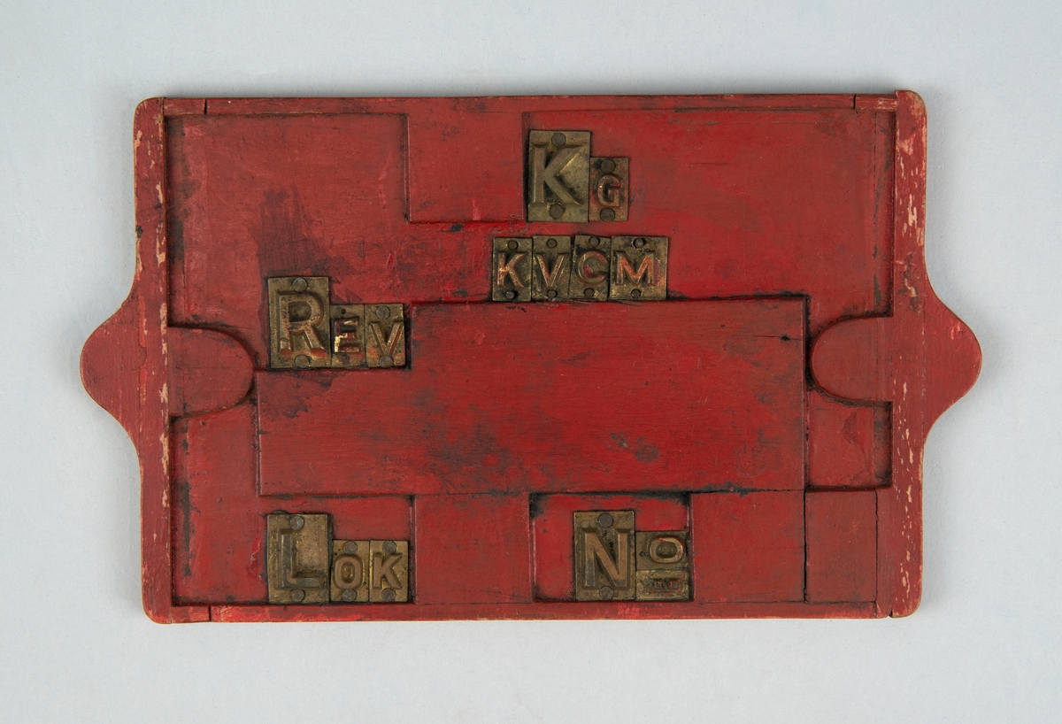 Rektangulär gjutförlaga till revisionsskylt av rödmålat trä. Metallbokstäver: "KG KVCM REV LOK No". På baksidan står det inpressat "1 - 353" samt en blyertsteckning.