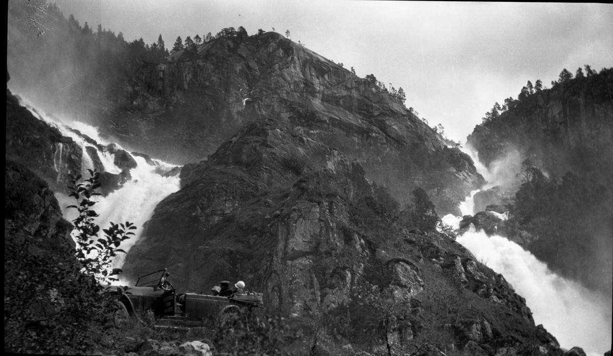 Fotosamling etter Kjetil Skomdal (1881-1971). Fotografen virket i perioden 1910 til 1960 og hadde eget mørkerom hvor han også tok imot film fra andre fotografer. I perioden var han også postbud i Fyresdal. Portrett med bil og folk foran fjell og fossefall.