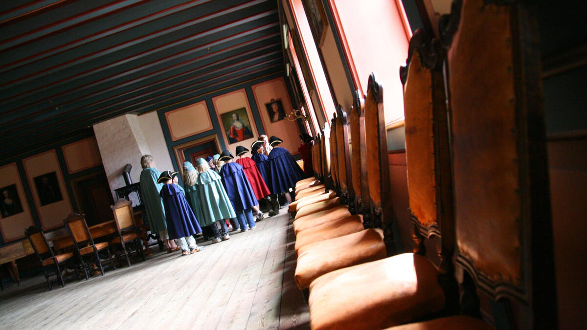 Bildet viser et rom i Herregården med stoler langs veggen. Innerst i rommet er en gruppe barn samlet, utkledd som grever og grevinner.