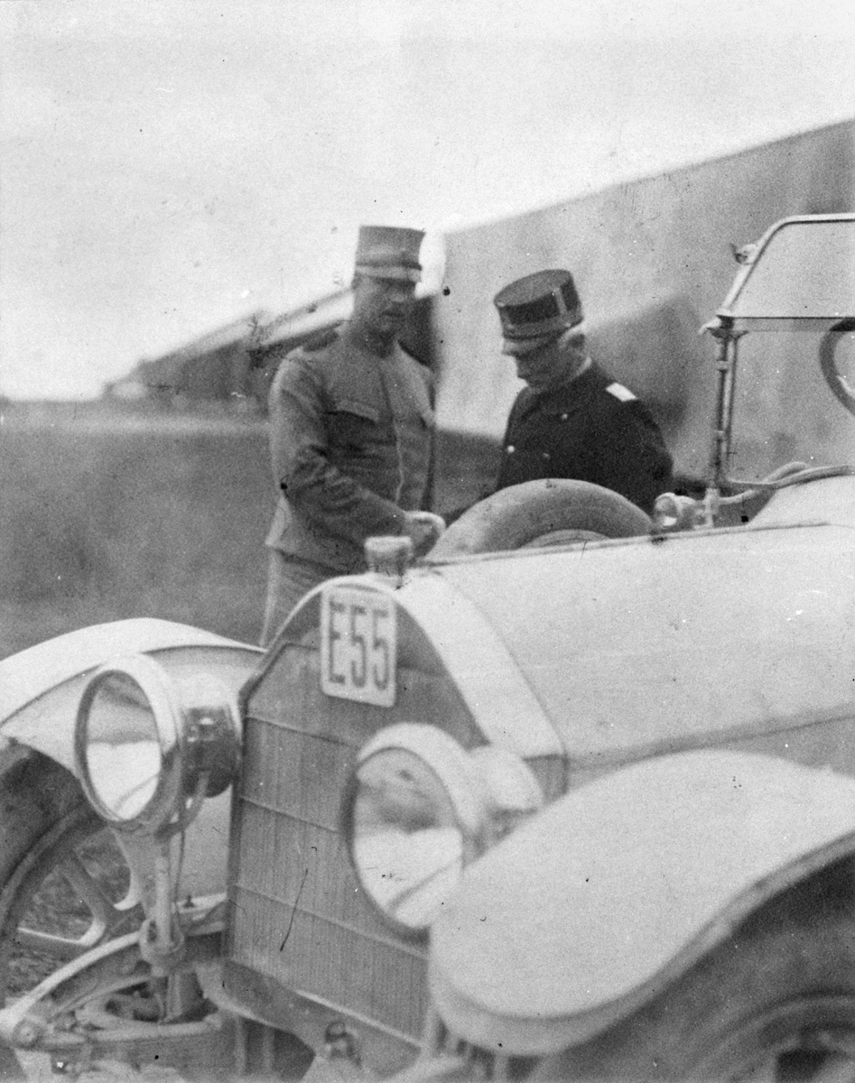 Två militärer står vid Flygkompaniets bil på Malmen, omkring 1917-1920. AEG- och F-hangarerna i bakgrunden. Till vänster Flygkompaniets chef Ernst Fogman.
