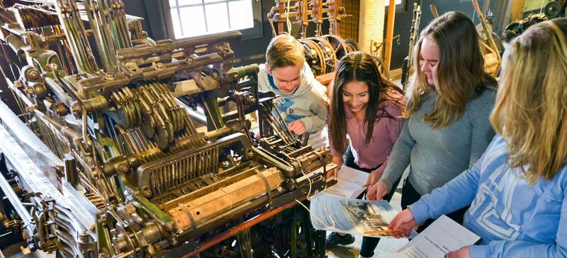 Bildet viser fire unge jenter som holder i penn og papir  ved siden av en maskin. (Foto/Photo)