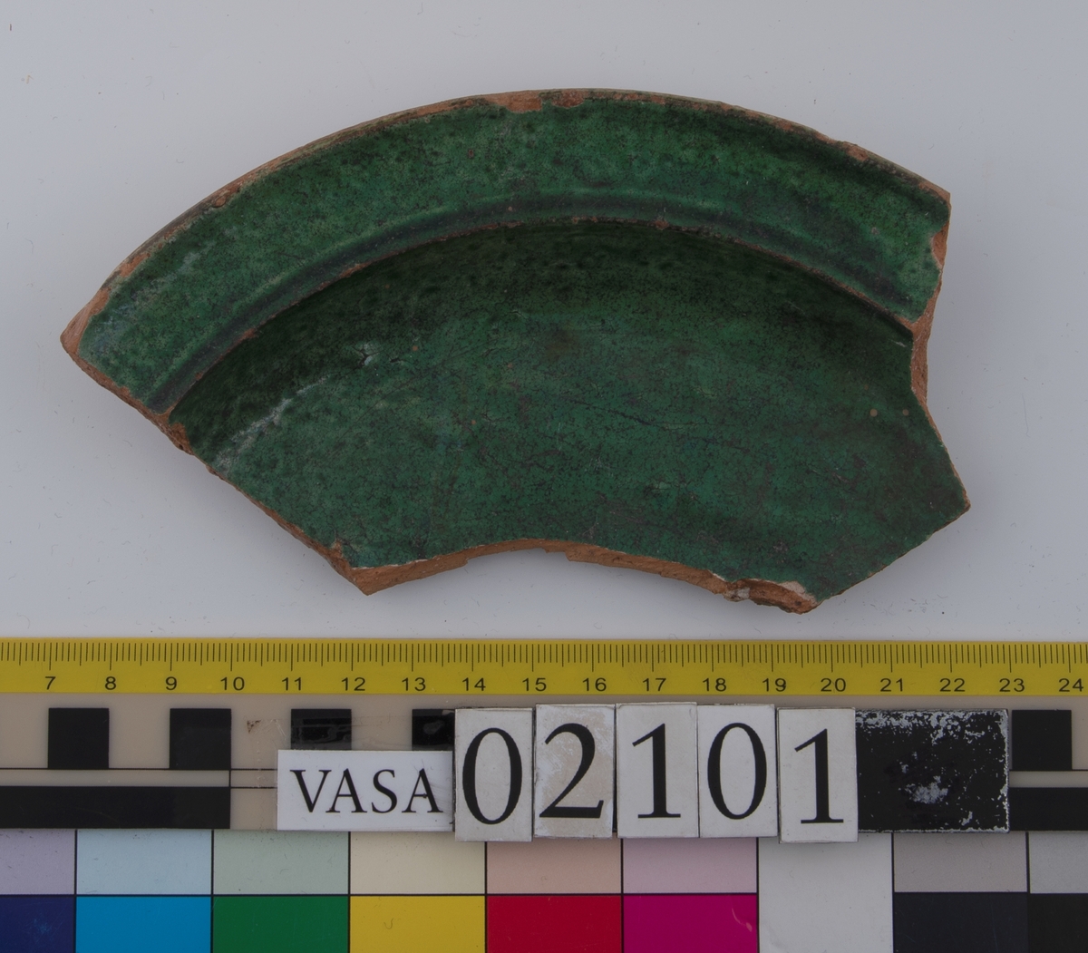 Två skärvor från ett grönglaserat keramikfat.
Fragment av mynningsrand (bräm) med bit av plan botten av fat. Profilerad bräm. Insida blyglaserad grön. Utsida oglaserad. Undersidan av den platta botten är räfflad.