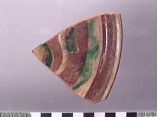 En mynningsskärva från en keramikskål. Skärvan är glaserad med en transparent glasyr och dekorerad. Dekoren består av våglinjer i grön och vit glasyr. På baksidan, närmast mynningskanten, löper tre drejfåror.