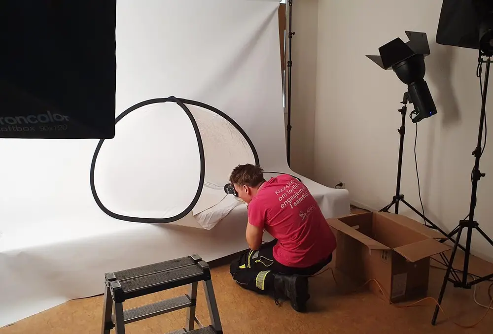 En mann i en rød t-skjorte i et fotostudio, foran han er et hvitt læret og en reflektor, han tar et fotografi av en gjenstand ute av syne