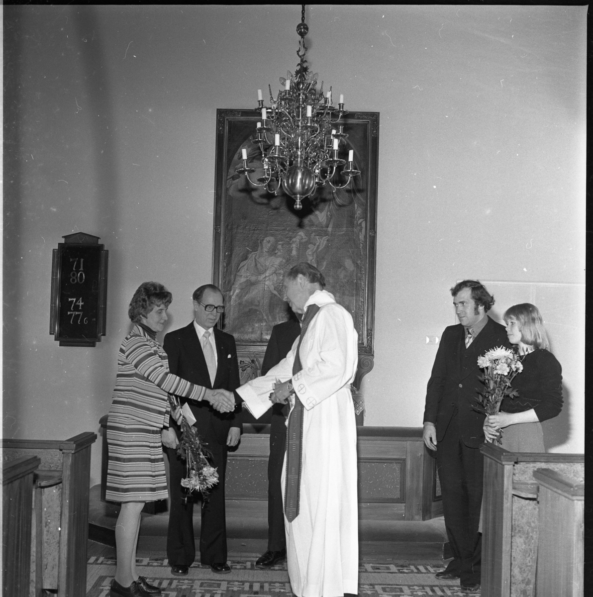 Män och kvinnor med blommor vid koret i Örserums kyrka. Det är avtackning och skifte av kyrkvaktmästare. Prästen handhälsar på en kvinna.