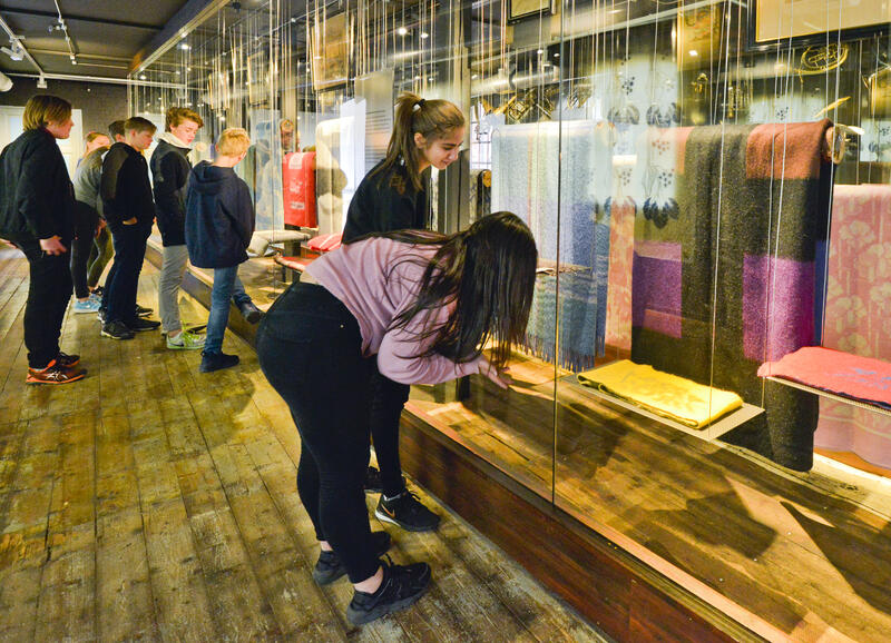 Bildet viser ungdommer som ser på en utstilling av tepper som henger i et avlangt stort glassmonter (Foto/Photo)