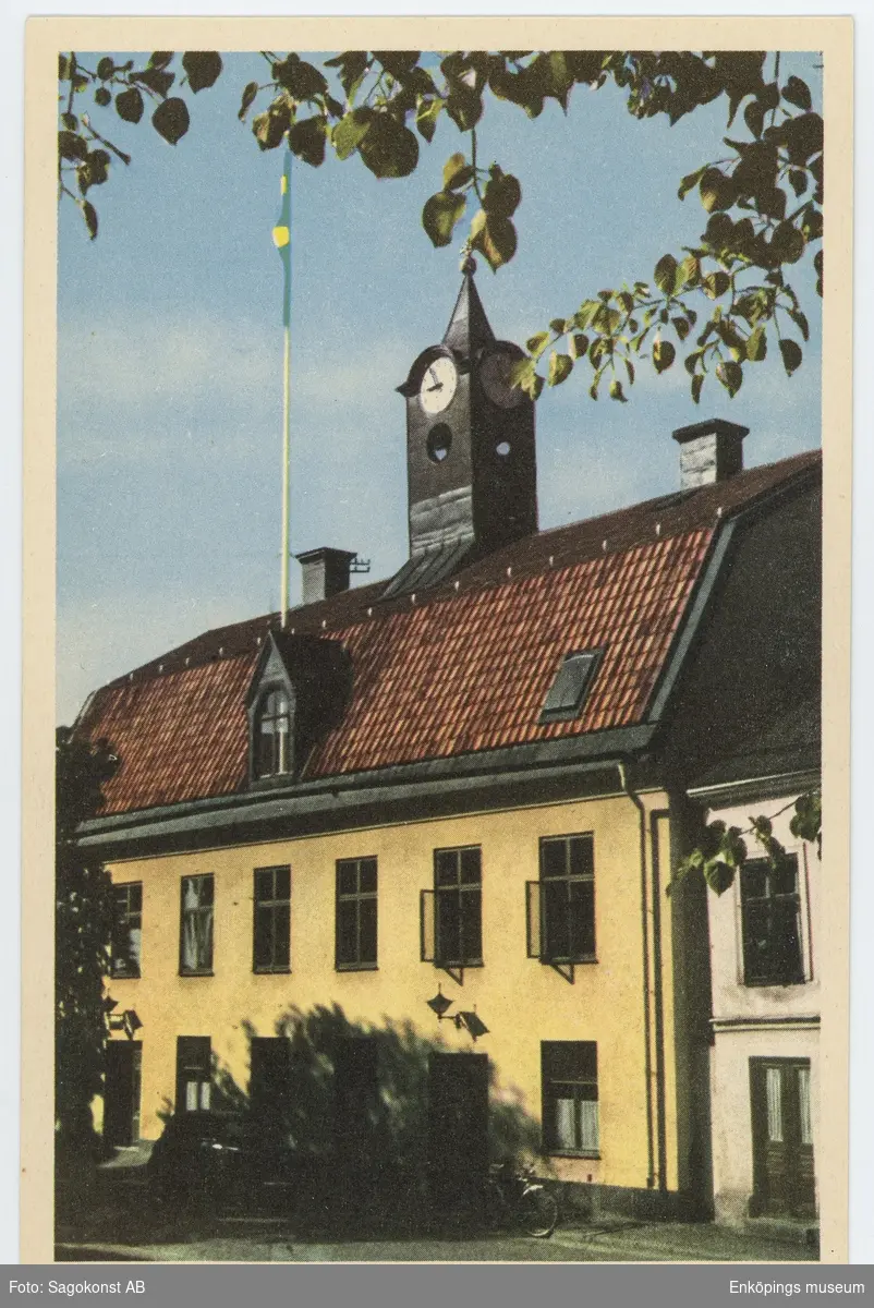 Vykort med motiv från Rådhusgatan, Enköping. Bilden visar det gamla Rådhuset som Nils Svensson Stahre lät bygga på Rådhusgatan 3 år 1783. Bygget tog 3 år och det byggdes som bostad och köpmannagård. Huset stod kvar efter den stora stadsbranden 1799 och i början av 1880-talet blev det rådhus. Senare blev det polishiús och idag huserar Enköpings museum och Turistinformation i byggnaden. Enköpings museum öppnade portarna i december 1987 och Turistinformationen flyttade in våren 2012.