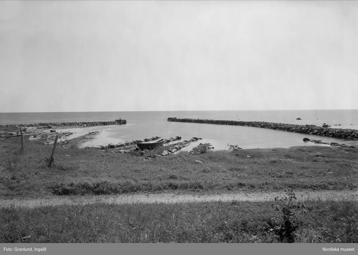 Havsstrand med vågbrytare av stenar, en båt ligger i vattenkanten. Burs socken, Gotland, Herta fiskeläge.