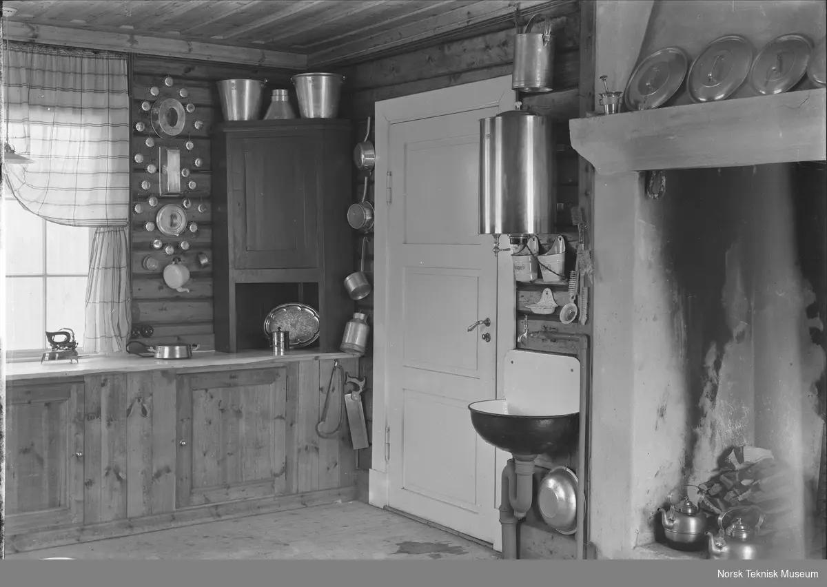 Mønsterkjøkken med elektrisk varmtvannsbeholder, kokeplate og strykejern, peis med vedstabel i forgrunnen, forskjellige kjøkkenredskaper henger på veggene : kjøkkenet er del av mønsterbruket på NEBB's jubileumsutstilling i 1914