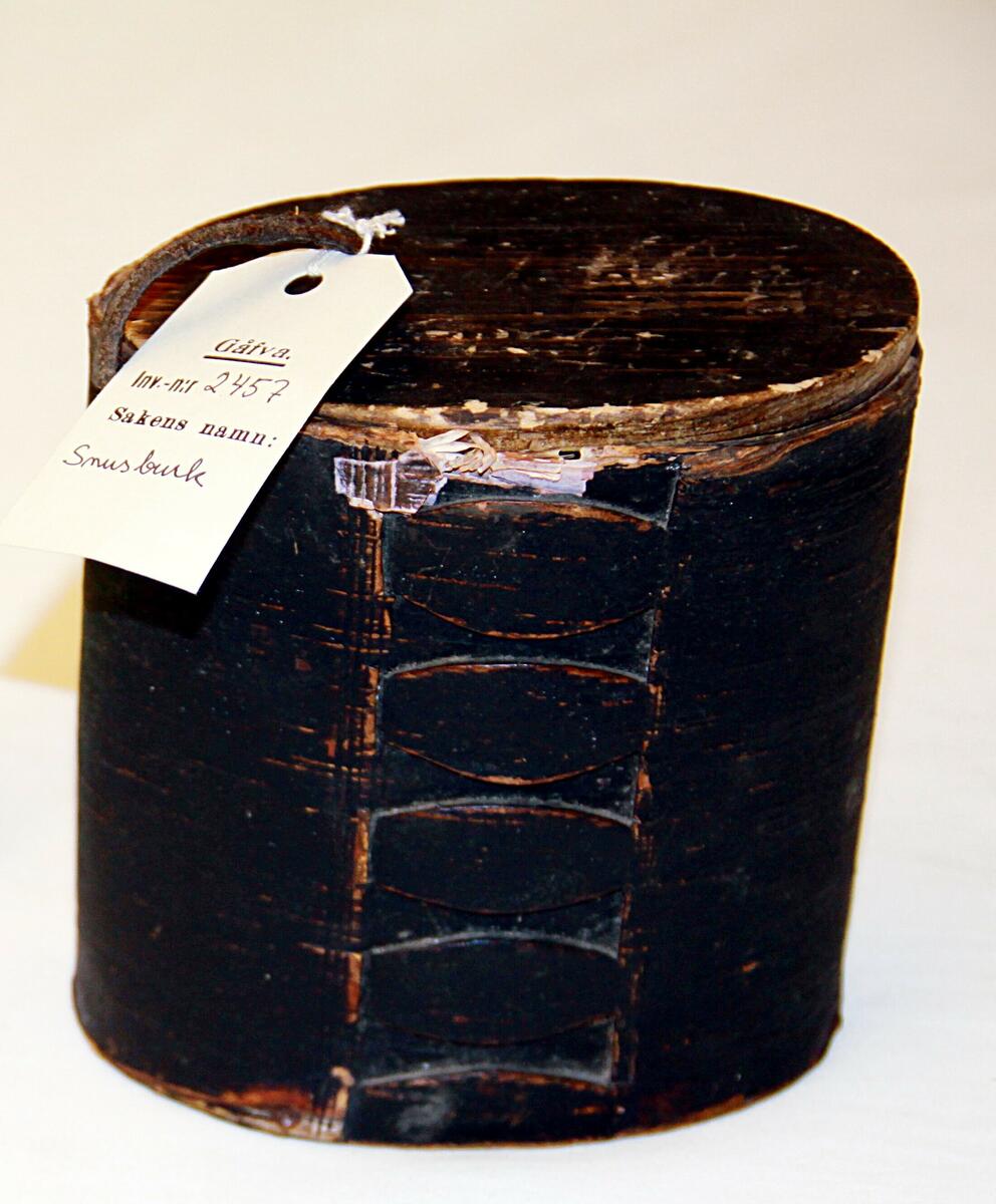 Snusburk av näver, svart-blåmålad på översidan med läderstropp, oval med infälld botten av trä lock av trä med läderband . Tillverkad 1895.
