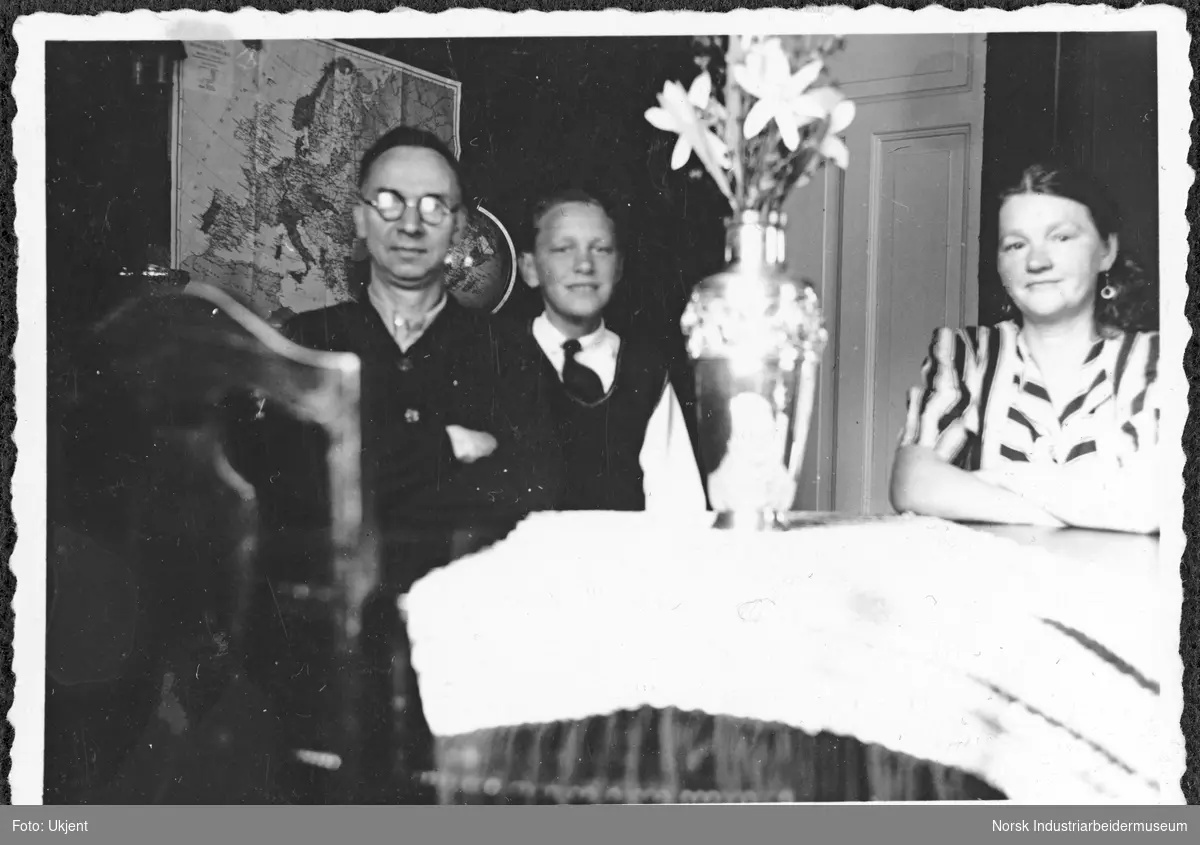 Mennesker sittende i stue rundt bord. Gustav Johansen var vaktmann på Vemork under operasjon Gunnerside og har på seg brillene som ble funnet av sabotørene under sabotasjeaksjonen.