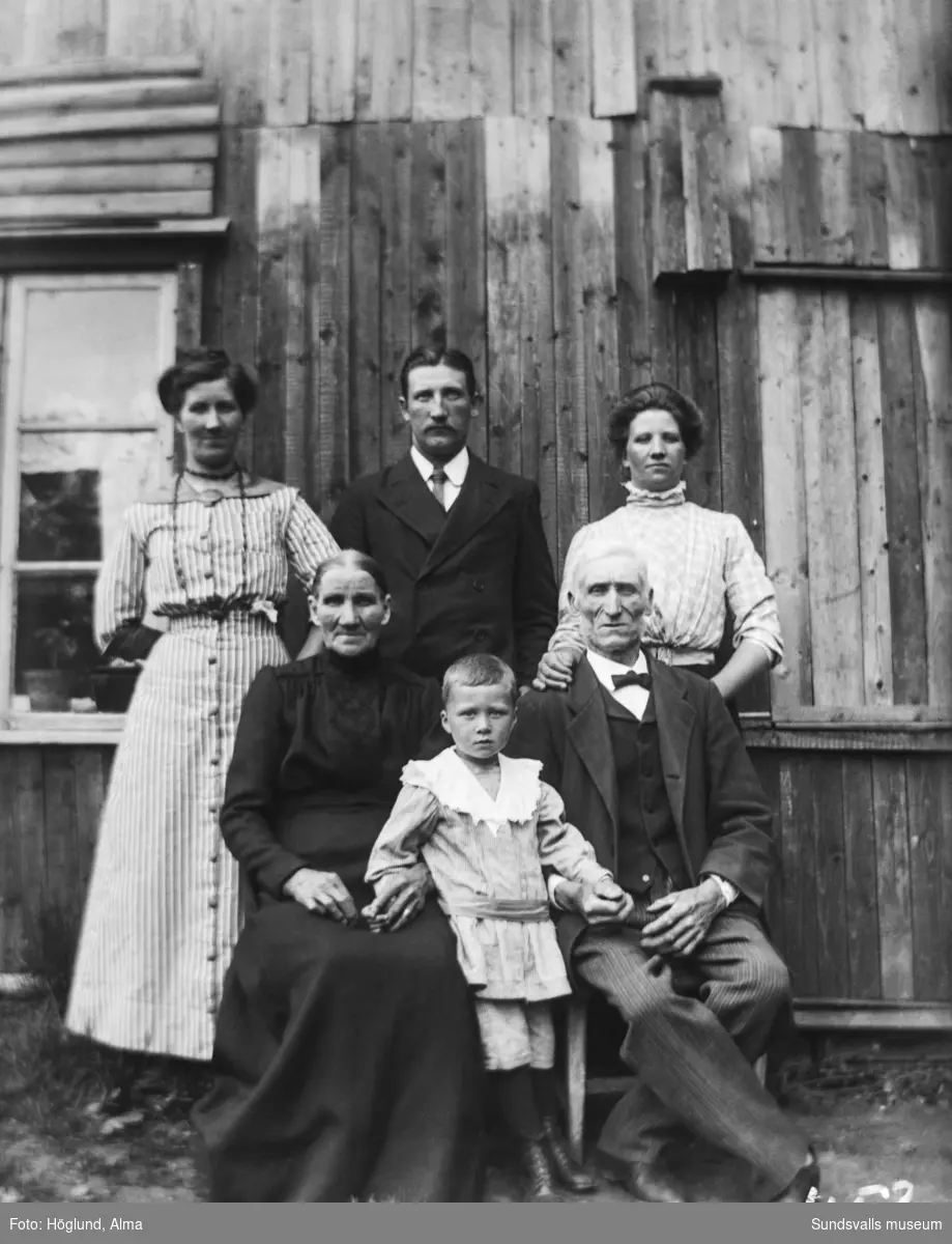 Familjebild med ett äldre par, två kvinnor, en man och en liten pojke invid en husvägg.