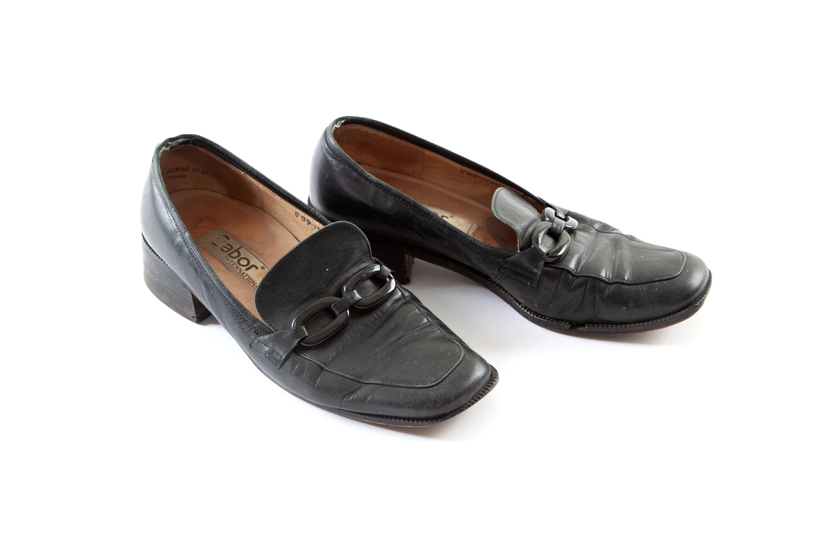 Et par svarte sko med lav/moderat hæl og pløse pyntet med svarte plastringer.