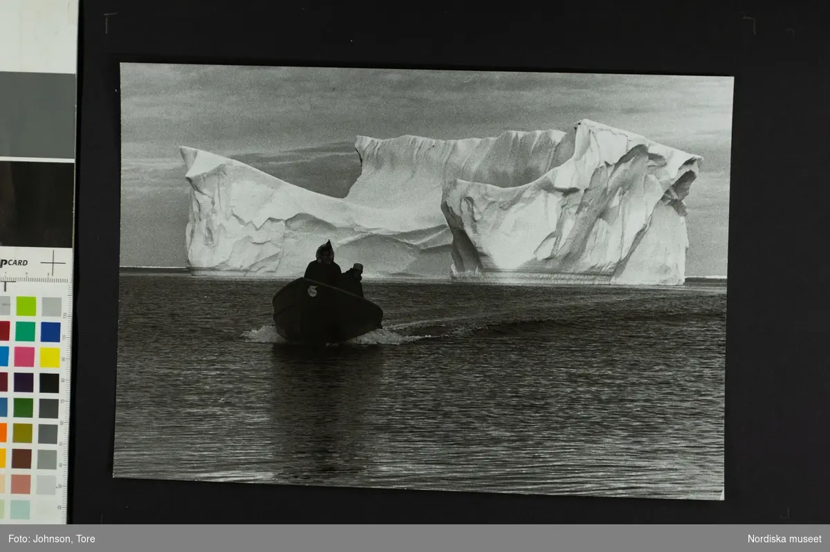 Två personer kommer åkande i en båt. Ett stort isberg på vattnet bakom dem. Mellan Baffin Island, Canada och Grönland, skolskjuts.