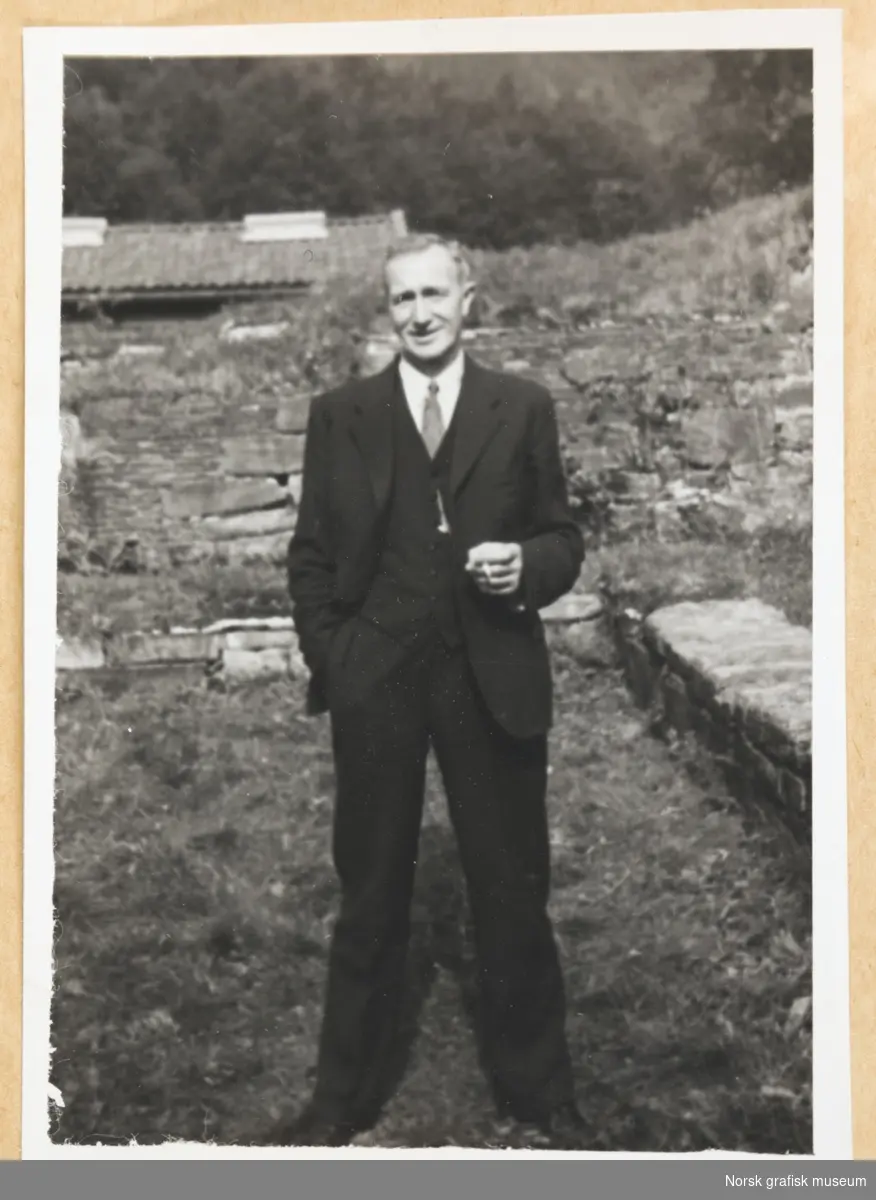 Utendørsmotiv. En mann står foran noe som ser ut som en gammel mur. I bakgrunnen skimtes et hustak med takpanner. Mannen er kledt i dress og slips, og holder en sigarett i venstre hånd. Fotografert i forbindelse med Vestlandsk Trykkerstevne i Bergen, 1946.