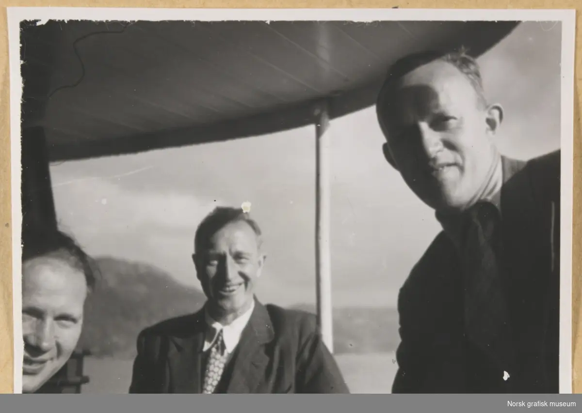 Utendørs under en slags overbygning, antatt ombord på en båt. Kystlandskap i bakgrunnen. Fotografert i forbindelse med Vestlandsk Trykkerstevne i Bergen, 1946.