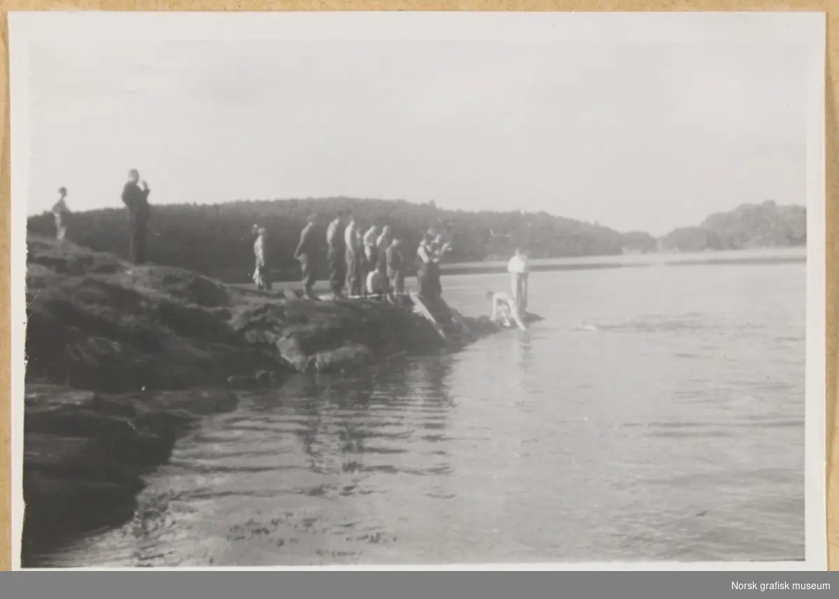 En gruppe mennesker i et kystlandskap. Noen bader i sjøen, men de fleste står påkledd på land og ser på. Fotografert i forbindelse med Vestlandsk Trykkerstevne i Bergen, 1946.