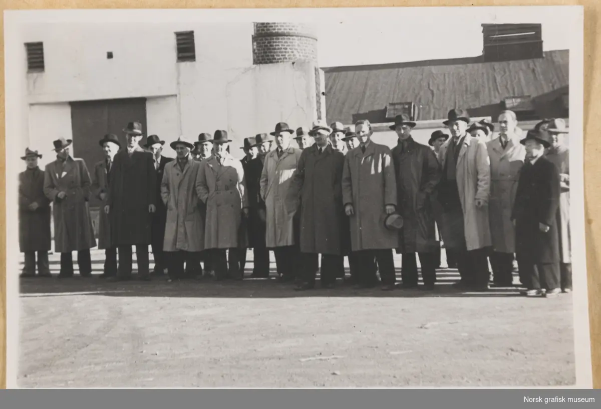 Utendørs gruppebilde. Rundt tjuefem menn i frakk og hatt står oppstilt foran en murbygning. Det skimtes noe som ser ut som en fabrikkpipe i murstein. Fotografert i forbindelse med Vestlandsk Trykkerstevne i Stavanger, 1948.