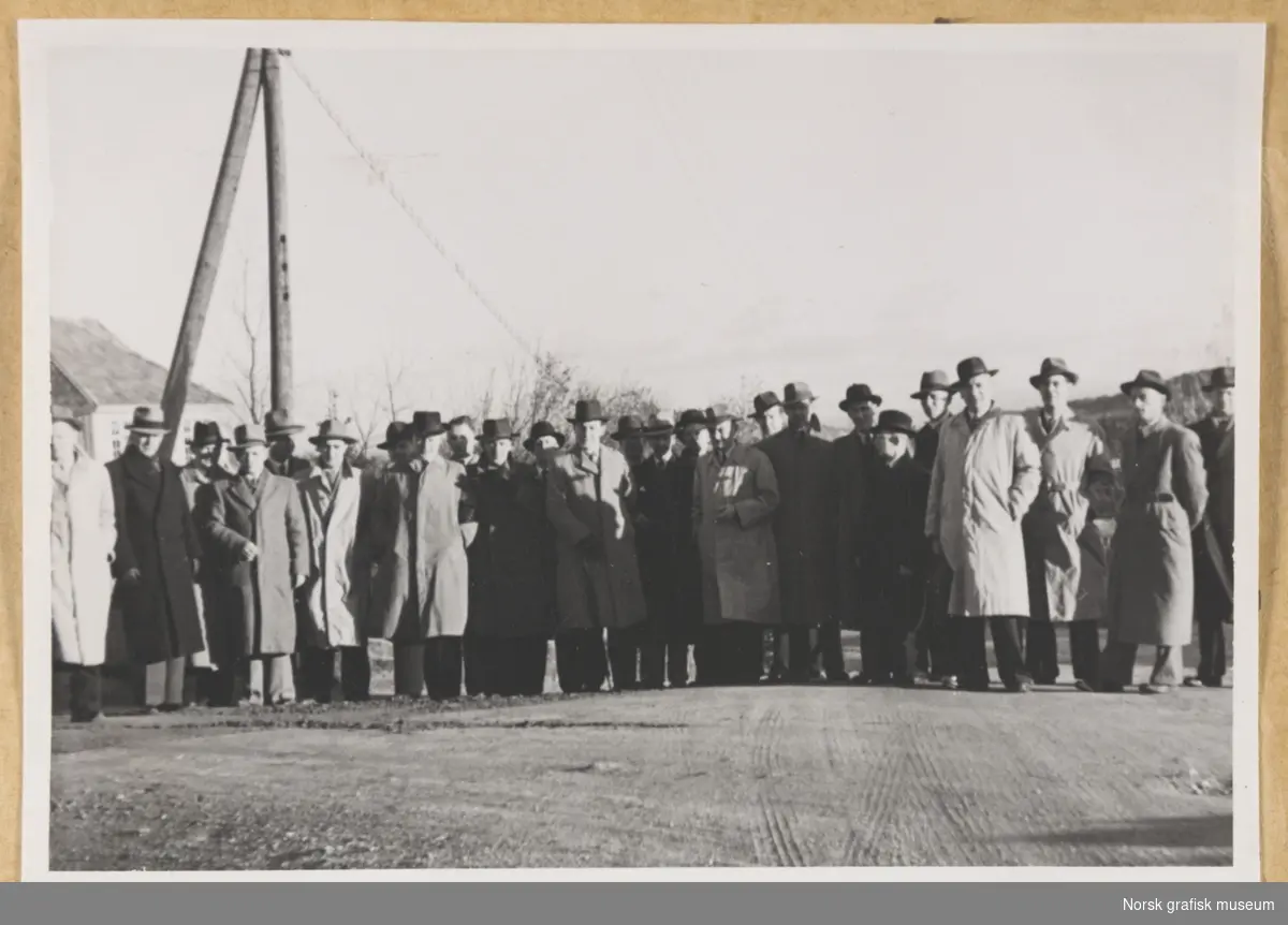 Utendørs gruppebilde. Rundt tjuefem menn i hatt og frakk står oppstilt på en grusdekt plass. Til venstre i bakgrunnen ser vi en lyktestolpe eller lignende, og toppen av et hus. Fotografert i forbindelse med Vestlandsk Trykkerstevne i Stavanger, 1948.