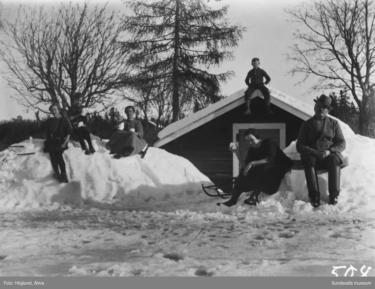 Hemma hos Höglunds poserar barn och vuxna i snödrivorna framför källarsvalen på gården i Fanbyn. På taknocken sitter sonen Per, framför från vänster Ingrid, Irma, Ada Wiström, Hilma och en oidentifierad man.