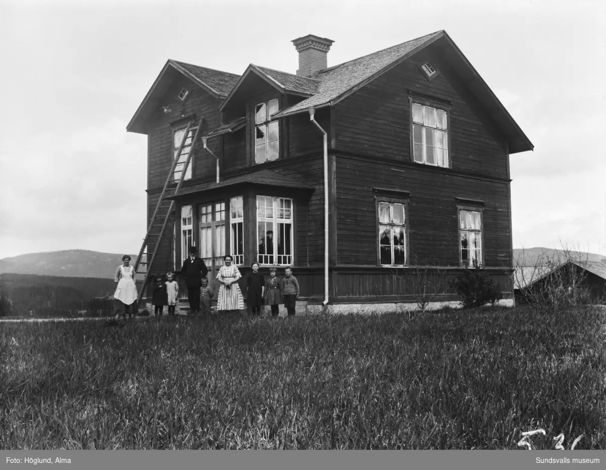 Två bilder på en familj framför glasverandan till sin bostad. En man, två kvinnor och sex barn. Andra bilden, en närbild på familjen.