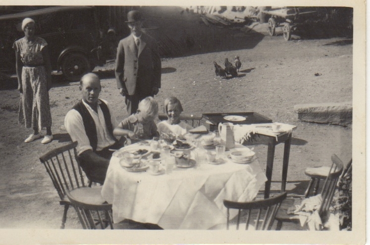 Axel Johansson (1895-1974) och sonen Bengt (1930-2016) sitter och fikar vid matbordet i trädgården, Labacka 1:2 "Lund" okänt årtal. Övriga personer är troligtvis sommargäster. I bakgrunden pickar några höns på marken.