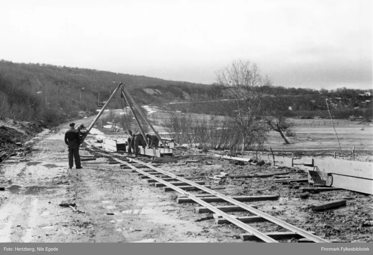 Tana bru ble sprengt november 1944. Sommeren 1948 ble Tana bru bygd på nytt. Flere menn jobber med å klargjøre en langbærer for transport. Oppsynsmann Edvin Eliassen til venstre.